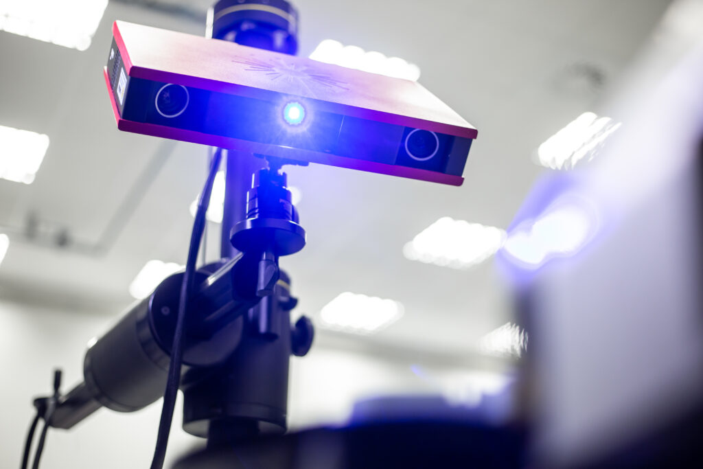 "Close-up van een professionele 3D-scanner op statief, met blauwe projectielichtstraal in een verlichte ruimte.